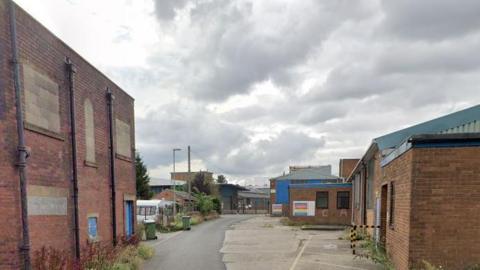 One and two-storey red brick industrial buildings line a partially tarmac-ed street which has weeds sprouting at the sides.