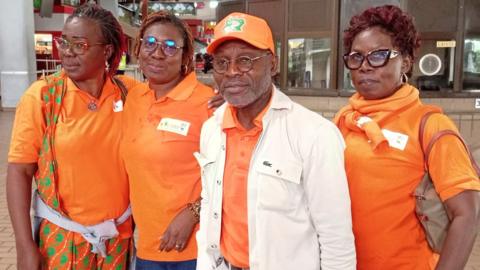 A man wearing an orange shirt and baseball cap and a white jacket looks at the camera. He has glasses and short facial hair. Three women stand with him, also wearing orange polo shits. 