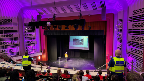 Interior of a theatre, showing a person in a yellow shirt and dark trousers walking on the stage near a small lectern, and an image projected on the back of the stage.  There are red curtains, and boxes to both sides with a wavy line pattern on the walls. Children are sat in the theatre seats, with a wooden shelf in front of them.  There are two police officer in yellow hi-viz standing up at the front of the seating area.