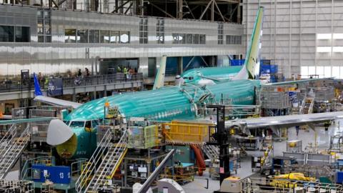 A large hanger in which a green aircraft with white wings is being built (a 737 MAX). There are railed access steps in several places. A mezzanine office area is visible to the left.