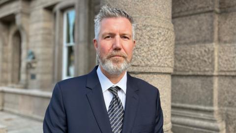 Adrian Moss is pictured in Jersey's Royal Square. He has a navy blazer on and a stripey tie. His hair is short, grey and spiky. He has a medium length beard. 