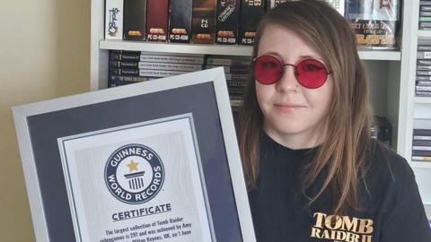 A woman wearing red Lara Croft sunglasses and a T-shirt with "Tom Raider 2" written on it is holding a frames Guinness World Record. She is stood in front of shelves stacked with video game boxes.