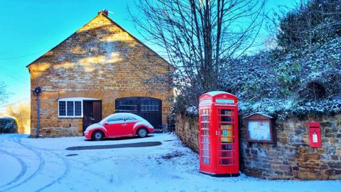 A sandstone how with a brown wood door and brown wooden garage doors. In front of it is a red VW Beatle car. to the right is a stone wall, red phone box and red post box embedded in the wall. The wall is in front of a bank with bushes and tree. The ground and foliage is covered in a light dusting of snow.