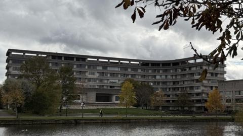 A large, curved concrete building with multiple rows of windows stands behind a grassy area and a pond. The building is Bedford Borough Council's headquarters. Several trees with autumn foliage are scattered across the scene, and branches frame the top of the image. The sky is overcast, creating a moody atmosphere, and a few people can be seen walking along the path near the water.