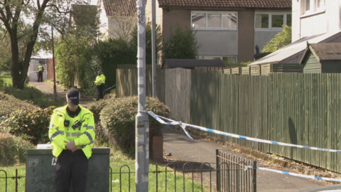 A footpath by some modern houses. There are trees and a grassy area on the left. Police tape has sealed off the path and three officers in hi-vis yellow vests can be seen at stages into the distance. It is sunny.