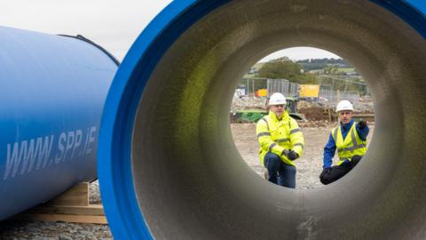 Two men pictured standing on other end of pipe, in high visibility jackets and hard hats. The picture is taken through a large pipe. It is blue. 