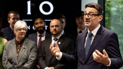 Burnham speaks in front of a crowd of people wearing a dark blue suit, a white shirt, a blue tie and black spectacles. He is gesturing and the audience of civil servants is listening intently. A large black sign bears an illuminated No 10 North label, written in the same font as No 10 on the famous black Downing Street door of the prime minister's official residence in London.