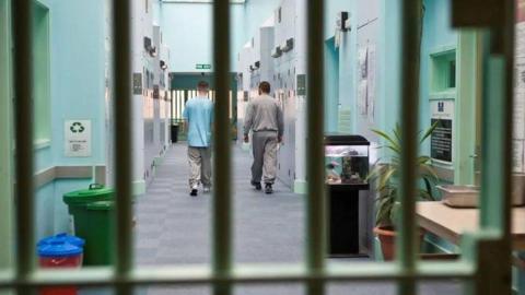 Two men walk away from us down a prison corridor with cells on either side. They have been pictured through the bars of a door.