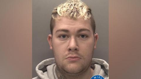 A mugshot of a man with dyed blond hair. He is wearing a grey hoodie and has a tattoo just visible on his neck. He also has a silver stud earring in the top of his left ear.