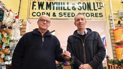 Peter Hodges is wearing a dark fleece jumper and standing on the left. Roy Hagan is on the right wearing a fleece-type cardigan on top of a dark jumper and shirt. They are standing in a shop. Behind them we can see the 'F Myhill and Son Ltd, Corn and Seed Stores' sign.