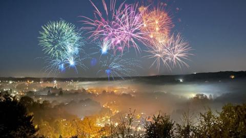 Fireworks over homes in a dark sky. The fireworks create bursts of different colours, green, blue, pink and reds, and underneath, slightly hazy from the smoke, are homes illuminated by domestic and street lights.