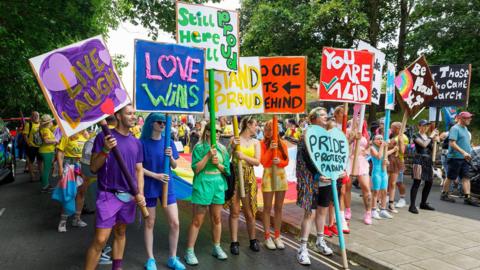 People dressed up in monochromatic colours of the rainbow holding up signs with their corresponding colours. The signs read phrases like "love wins" and "you are valid"