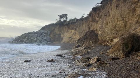 A windswept beach scene with eroding sandy cliffs, pebbles, waves and granite rock defences in the background 