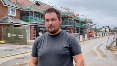 Nathan Long stand on the street in front of a houses with scaffolding and builder's fencing.