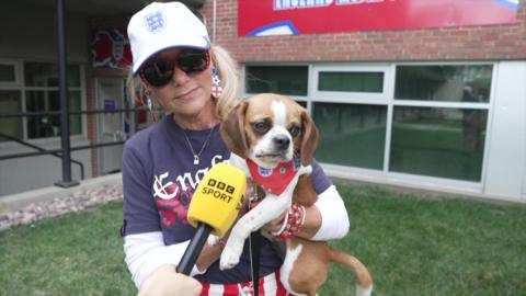 A blonde lady wearing an England shirt and cap holds a dog wearing a red England bandana