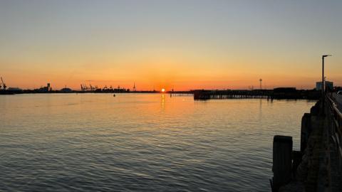 MONDAY - The sun sets over Southampton docks, on the horizon the sun appears as a small orange ball and the sky is glowing orange, you can see the silhouettes of cranes. In the foreground is the water in the harbour with the sun reflected in the ripples. There is a pier stretching across the middle of the picture from right to left.