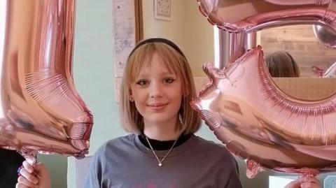 A teenage girl with shoulder-length blonde hair wearing a silver necklace and grey jumper smiling into the camera. She is standing in a room of the house holding up two pink balloons. One is in the shape of the number 1 and the other is in the shape of the number 5.