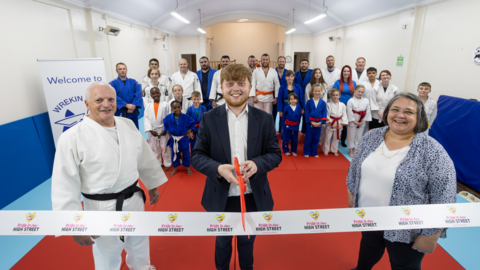 Councillor Ollie Vickers, centre, has short brown hair and is wearing a dark suit with a white shirt. He us holding a giant pair of scissors ready to cut a white ribbon in front of him. He is stood next to Anna Halliday, who has bobbed grey hair and is wearing a blue and white shirt, white T-shirt and dark trousers, and Gareth Mapp, who has white hair and is wearing a white judo uniform with black belt. They are at the opening of Wrekin Star Judo.