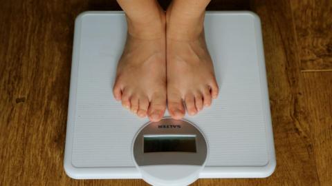 A person stands on a pair of white digital weight scales, only their bare feet can be seen.