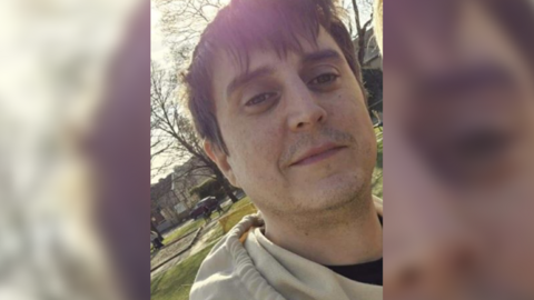 Benjamin Swift, a man in his late 30s, looks at the camera. He has short dark hair and dark eyebrows. He is wearing a cream coloured hoodie. A suburban street is visible in the background.