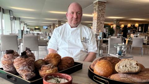 A person wearing a white chef’s jacket sits at a table in a spacious, modern restaurant, with trays of pastries and assorted breads arranged in the foreground.