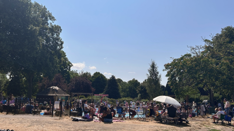A crowd of people, mainly families, inside and surrounding the paddling pool. Some have parasols to protect from the sun. There is grass in the foreground and trees in distance