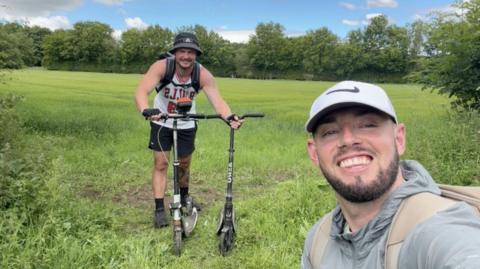 A man holding two black push scooters while standing in a field. He is wearing a backpack, a black bucket hat, a white vest, black shorts and trainers. In front of him taking the selfie is a man wearing a white Nike-tick hat and a grey coat, with a beige backpack. They are standingd in a field.