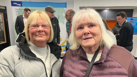 Two women in padded jackets pose together at a busy indoor event, with other attendees and exhibition displays behind them.