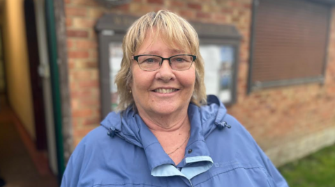 A woman pictured outside a building wearing reading glasses and a blue raincoat. She has short blonde hair and is smiling.