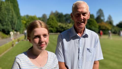 Emily is on the left. She has blonde hair which is tied back in a ponytail and is wearing a stripy top and a necklace. Her grandad is standing next to her. He is tall, has white hair and is wearing a white and blue checked shirt.