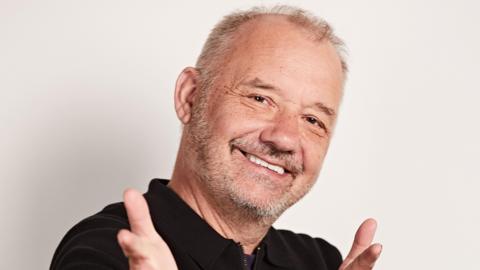 Bob Mortimer with white short hair and a grey stubble beard, holding his hands up, wearing a black fleece. 