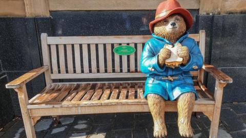 Statue of Paddington Bear sitting on a bench eating a sandwich.