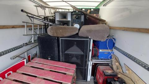 The inside of a  white van with lots of clutter including a table, poles, cushions and large speakers. The objects are piled on top of each other. 