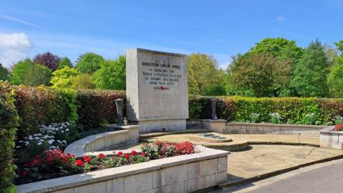 A stone memorial area. The memorial is surrounded by leafy green trees and hedges with red and white flowers planted in front of a walled area. An inscription on the memorial reads "In memory of those citizens killed in enemy air raids over this city".
