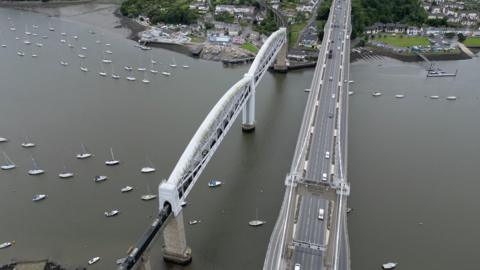 An aerial view of the Tamar Bridge road crossing between Cornwall and Devon with boats in the river.
The road appears almost empty but a few vehicles ae driving in either direction.  Houses and tress in Saltash in Cornwall also appear in the image.
Isambard Kingdom Brunel's railway bridge with two arches is in the left of the picture.