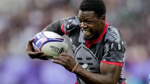 Vincent Onyala, wearing a grey Kenya shirt with red trim, grimaces as he runs forward holding a rugby ball in his right hand