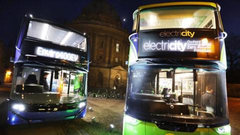 The new OBC and Stagecoach electric buses at the Oxford launch. The two busses - one purple and one green - are parked in front of the Radcliffe Camera which can be seen in the background. There are bicycles in front of the building. The picture is taken during night time and the busses' lights are on.