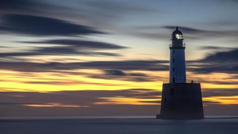 A lighthouse in the sea. The sky is orange with dark clouds streaked across it.