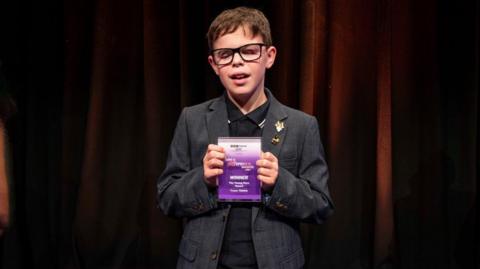 A young boy with short brown hair and wearing glasses and a dark grey suit. He is holding a purple and white award in both hands. Behind him is a dark red or brown curtain. 