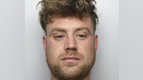 Close crop of a police mugshot. The man has a wide lower face and large eyes. He is looking down, slightly past the camera. He has a light brown beard.