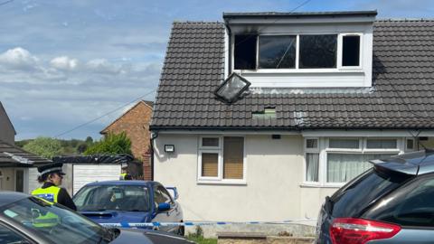 House following fire with a burnt-out window and a police officer standing outside