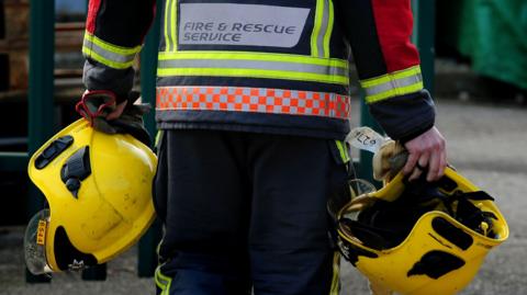 A firefighter on a training session. He is seen carrying two yellow helmets. He wears a long sleeved top with Fire and Rescue Service sign on the back. He has his back to the camera.