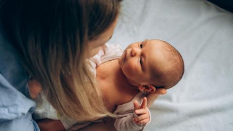 A mum, who is wearing a blue shirt and has shoulder length fair hair, leans over while holding her baby. The baby is looking up into her face.