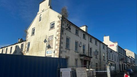 The Antrim Arms Hotel, a white building. Antrim Arms is written in red letters above the door. There are bollards and a fence in front of it.