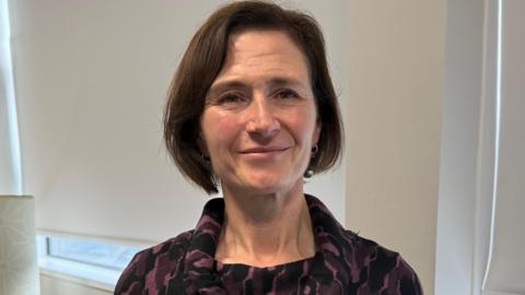 Annette Morris has short, brown hair wearing a dark purple and black patterned dress with a cowl neck in an office setting.