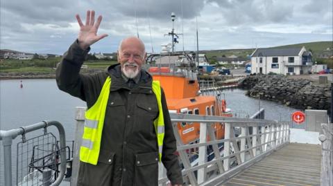 Derek Adams with short white hair and beard, wearing a dark green waterproof coat and yellow hi-vis jacket. He is waving with his right hand. Behind him is the orange RNLI Charles Lidbury lifeboat, moored alongside a landing stage with white railings. The sea is visible in the background.