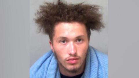 A police custody picture of a man with a blue towel or blanket around his neck.