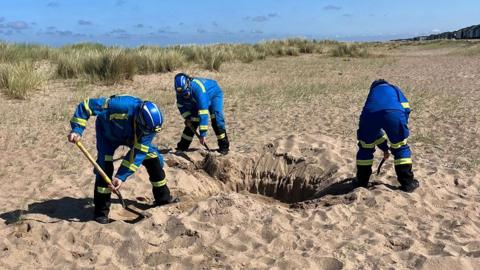 Three coastguards wearing blue protective clothing and helmets with yellow stripes use shovels to fill in a large hole on a sandy beach