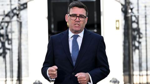 Andy Burnham speaks outside 10 Downing Street after becoming prime minister. He is wearing black glasses, a navy blue suit, white shirt, and blue cotton tie.