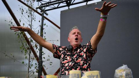 DJ Fatboy Slim plays on stage at Glastonbury Festival. He is stood in front of a darkened screen and has his hands in the air with his mouth open. He has short grey hair and is wearing a black shirt with orange and white koi fish on it. He has a pair of headphones around his neck.